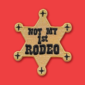 May include: A gold sheriff badge patch with the text "NOT MY 1st RODEO" in black. The badge is a six-pointed star shape with a cross at the end of each point. The background is a solid red.