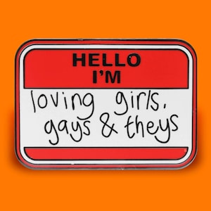 Peut inclure: Épinglette en émail rouge et blanc avec un design de badge. Le texte sur l'épingle est "Hello I'm loving girls, gays & theys".