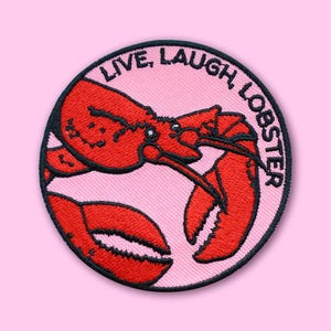 Può includere: Una toppa rotonda ricamata con un'illustrazione di aragosta rossa su sfondo rosa. La toppa presenta la scritta "LIVE, LAUGH, LOBSTER" in nero, circondata da un bordo nero. Adatta per abbigliamento o accessori.