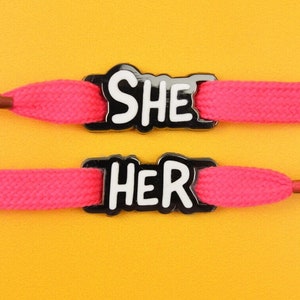Puede incluir: Dos pares de cordones rosa brillante con dijes en blanco y negro que dicen "SHE" y "HER". Los cordones están sobre un fondo amarillo brillante. Los cordones son de estilo plano y tejido.
