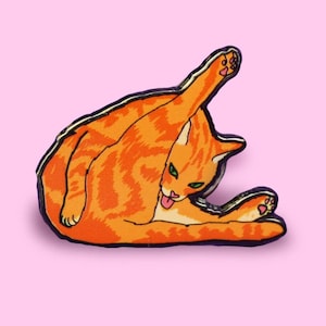 Può includere: Spilla a forma di gatto tabby arancione con occhi verdi e una lingua rosa che sporge. Il gatto è rannicchiato in una palla e si lecca la zampa.