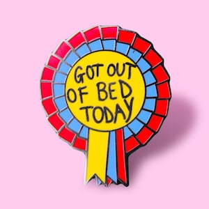 Puede incluir: Un pin de esmalte amarillo, rojo y azul con una cinta. El pin dice "Got out of bed today".