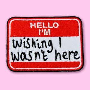 May include: Red and white name tag with black text that reads "Hello I'm Wishing I Wasn't Here".