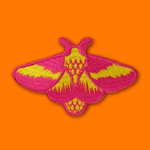 Puede incluir: Un parche de polilla bordado en rosa y amarillo. La polilla tiene un diseño detallado con un cuerpo amarillo y alas rosas.