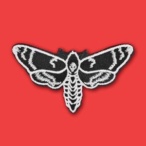 May include: Black and white embroidered patch of a moth with a skull-like face.