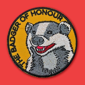 Puede incluir: Un parche bordado amarillo y negro con un tejón y el texto "The Badger of Honour".