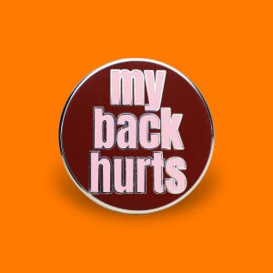 May include: A circular enamel pin with a deep maroon background and a silver metal border. The text "my back hurts" is written in bold, light pink capital letters across three lines, centered on the face of the round pin.