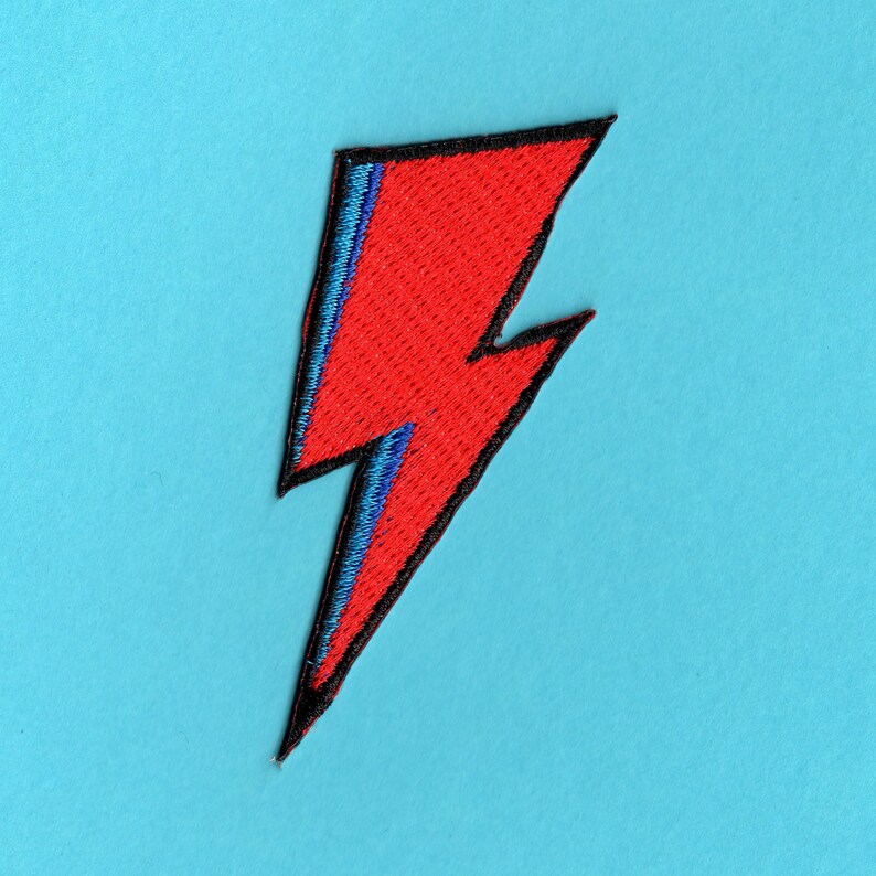Peut inclure: Un patch en forme d'&eacute;clair rouge et bleu avec des coutures noires. Le patch est sur un fond bleu clair.