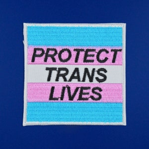 Pu&ograve; includere: Una toppa bianca con un design di bandiera transgender arcobaleno blu e rosa. Il testo "PROTECT TRANS LIVES" &egrave; ricamato in nero.