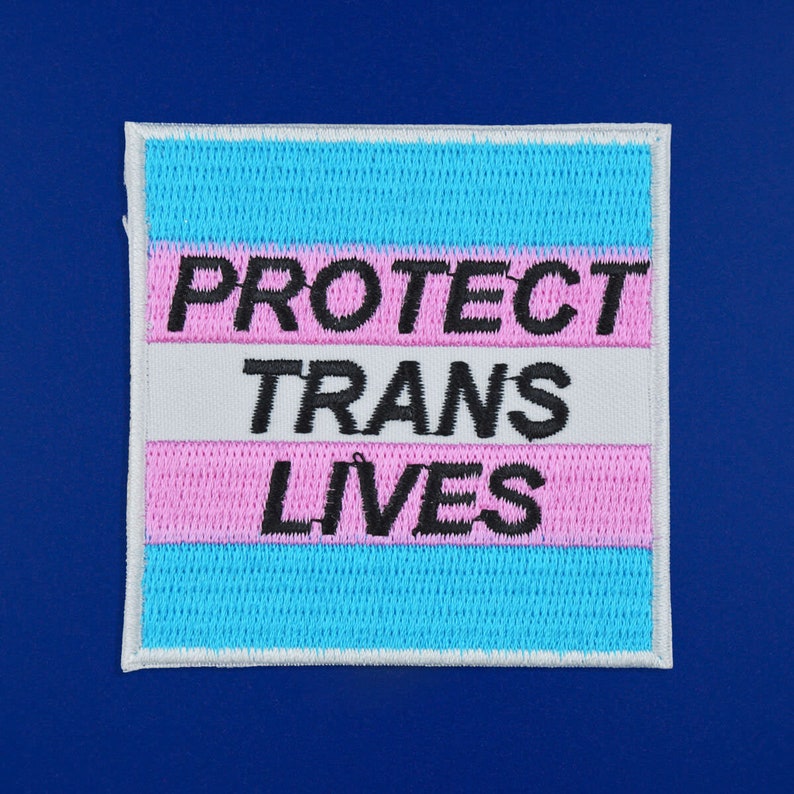 Pu&ograve; includere: Una toppa bianca con un design di bandiera transgender arcobaleno blu e rosa. Il testo "PROTECT TRANS LIVES" &egrave; ricamato in nero.