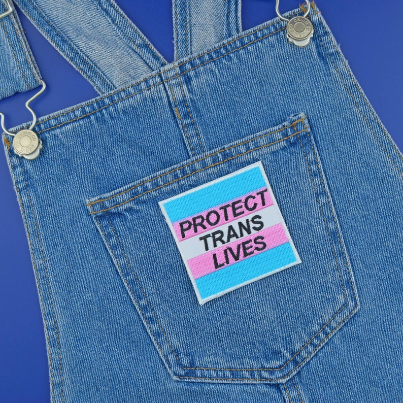 Pu&ograve; includere: Una salopette di jeans blu con una tasca che contiene una toppa bianca con il testo "Protect Trans Lives" in lettere nere. La toppa ha uno sfondo blu chiaro e rosa.