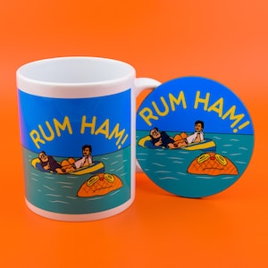 Może przedstawiać: Biały kubek ceramiczny z niebiesko-żółtą ilustracją trzech osób w tratwie na wodzie. Tekst "RUM HAM!" jest nadrukowany na kubku i podkładce w kolorze żółtym. Podkładka jest okrągła, niebieska z żółtą obwódką.