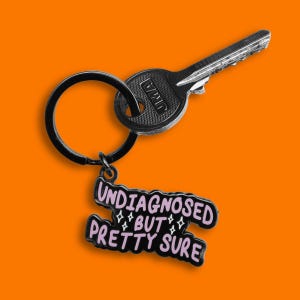 Può includere: Un portachiavi nero con una chiave d'argento e un ciondolo in smalto nero e rosa. Il ciondolo recita "UNDIAGNOSED BUT PRETTY SURE" in un font stilizzato con accenti di stelle. La chiave ha le lettere "JMA".