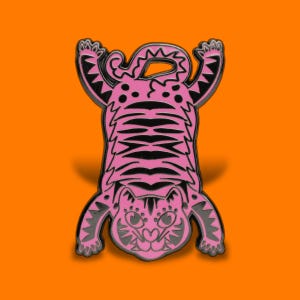 May include: A pink and black enamel pin shaped like a tiger viewed from above. The tiger is depicted with its limbs extended outwards, featuring black stripes across its body, a detailed face with large eyes, and a curled tail at the top.