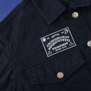 May include: A black denim jacket with a white embroidered patch featuring a classic Ouija board design. The patch includes the letters of the alphabet, numbers, and the words "Yes", "No", "Good Bye", and "Ouija".