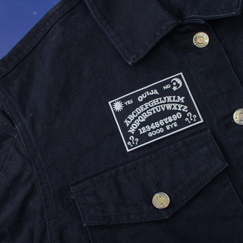 May include: A black denim jacket with a white embroidered patch featuring a classic Ouija board design. The patch includes the letters of the alphabet, numbers, and the words "Yes", "No", "Good Bye", and "Ouija".