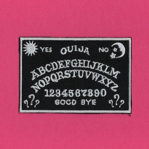 May include: Black and white embroidered patch with a Ouija board design. The patch features the letters of the alphabet, numbers 1-9 and 0, the words "Yes", "No", "Goodbye", and symbols of a sun and a crescent moon.
