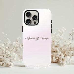 May include: A white phone case with a gradient pink and white background. The case features the text "Made in His Image" in a cursive font. The phone case has a black camera module and black side buttons.