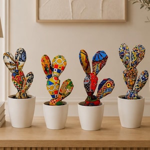 Puede incluir: Cuatro esculturas de cactus de tela en macetas de cerámica blanca. Cada cactus presenta múltiples paletas redondeadas cubiertas con tela de patrones multicolores vibrantes, incluyendo formas geométricas, motivos florales y estampados artísticos abstractos.
