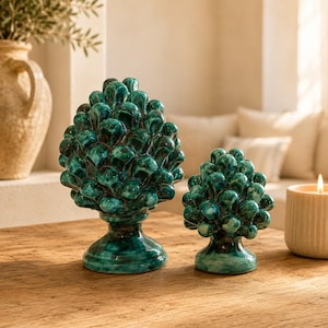 Può includere: Due sculture decorative in ceramica a forma di pigna, caratterizzate da una finitura lucida color verde ottanio con sfumature verde scuro. Ogni pezzo presenta squame arrotondate ed è montato su una base circolare, posizionato in verticale.
