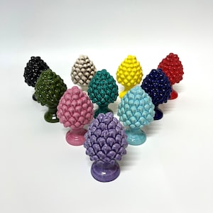 Può includere: Una collezione di sculture a forma di pigna in ceramica colorata, in vari colori tra cui nero, verde, rosa, viola, blu, giallo e rosso. Ogni pigna è posta su una base a piedistallo. Le sculture sono disposte su una superficie bianca.