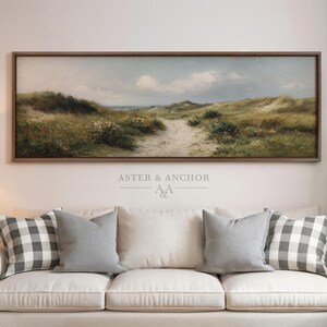 May include: A framed landscape painting depicts a sandy path winding through grassy dunes under a cloudy sky. The artwork is in a dark brown frame and hangs above a white sofa with gray and plaid pillows. The text "ASTER & ANCHOR" is visible.