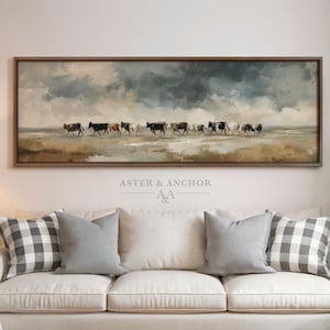 May include: A rectangular framed artwork depicts a herd of cattle walking across a field under a cloudy sky. The artwork is above a white sofa with gray and checkered pillows. The text "ASTER & ANCHOR" is in the lower right corner.