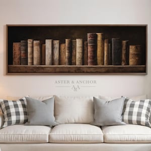 May include: A framed artwork featuring a collection of antique books on a shelf. The books have aged, textured covers in shades of brown and tan. The artwork is above a white sofa with gray and plaid pillows. The text "ASTER & ANCHOR" is visible.