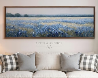 May include: A framed landscape painting depicts a field of blue flowers under a hazy sky. The artwork is mounted above a white sofa with gray and checkered pillows. The text "ASTER & ANCHOR" is visible in the lower right corner.