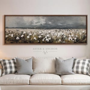 May include: A framed landscape painting depicts a field of white cotton flowers under a stormy gray sky. The artwork is above a white sofa with gray and white plaid and solid gray throw pillows. The text "ASTER & ANCHOR" is visible.