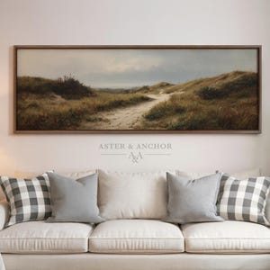May include: A framed landscape painting depicts a sandy path winding through grassy dunes under a cloudy sky. The artwork is in a dark wooden frame and hangs above a white sofa with gray and plaid pillows. The text "ASTER & ANCHOR" is visible.