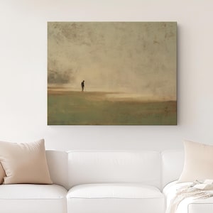 May include: Square canvas art print depicting a solitary figure on a golf course. The artwork features a muted color palette of greens, browns, and beige, creating a serene and contemplative atmosphere. The print is displayed above a white sofa.