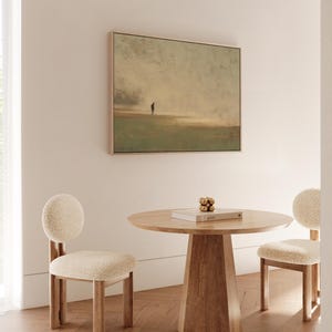 May include: A square framed artwork depicts a solitary figure on a muted landscape. A round wooden table with a book and decorative gold spheres is centered, flanked by two chairs with rounded backs and cream-colored upholstery. The room has a minimalist design.
