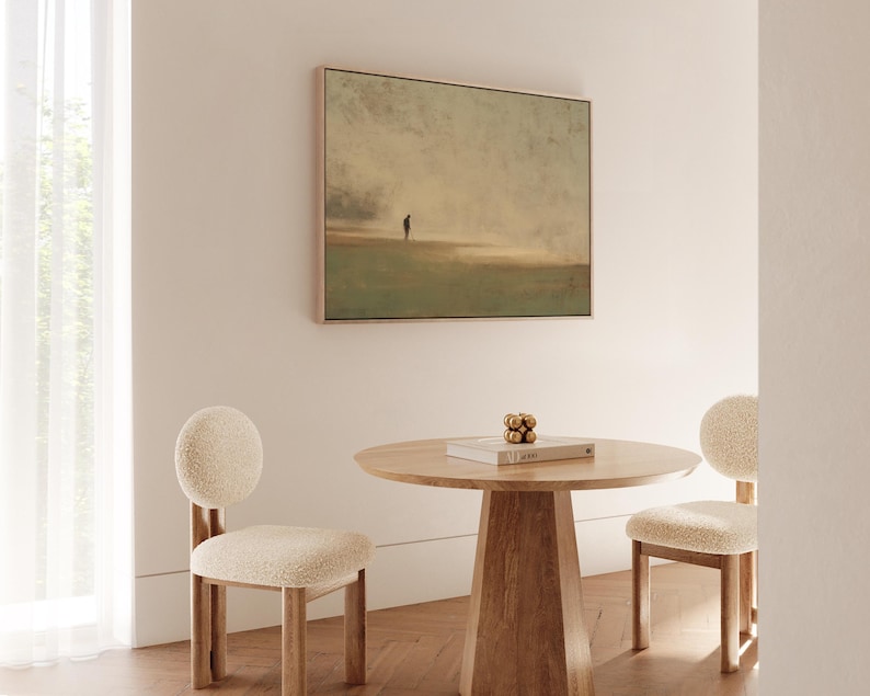 May include: A square framed artwork depicts a solitary figure on a muted landscape. A round wooden table with a book and decorative gold spheres is centered, flanked by two chairs with rounded backs and cream-colored upholstery. The room has a minimalist design.