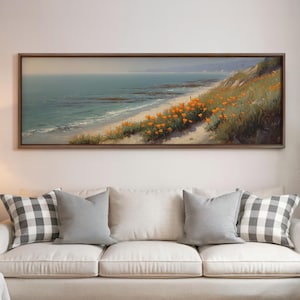 May include: A framed landscape painting depicts a coastal scene with a sandy beach, turquoise water, and a hillside covered in orange flowers. The artwork is displayed above a white sofa with gray pillows.