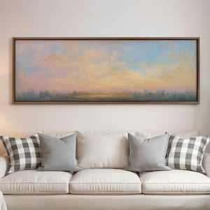May include: A framed landscape painting with a sky of pastel colors, including pink, yellow, and blue, hangs above a white sofa. The sofa has gray pillows and black and white checkered pillows.