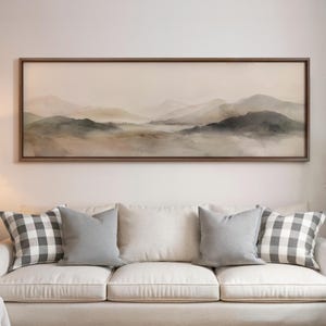 May include: A framed landscape painting depicts a mountain range in muted tones of beige, gray, and green. The artwork hangs above a white sofa with gray and checkered pillows, creating a serene living room scene.