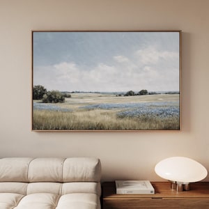 May include: A framed landscape painting depicts a field of wildflowers under a cloudy sky. The artwork features a palette of blues, greens, and browns, with a wooden frame. The painting is displayed on a neutral-colored wall.