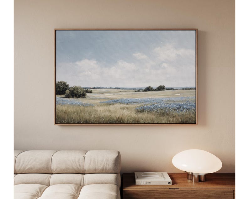 May include: A framed landscape painting depicts a field of wildflowers under a cloudy sky. The artwork features a palette of blues, greens, and browns, with a wooden frame. The painting is displayed on a neutral-colored wall.