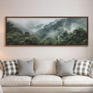 May include: A framed landscape art print depicting a misty mountain scene with lush green trees. The artwork is displayed above a cream-colored sofa with gray and checkered pillows. The frame is a dark brown color.