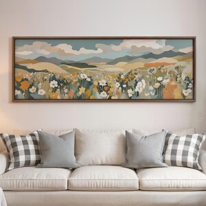 May include: A framed landscape art print depicting a field of wildflowers under a cloudy sky. The artwork features a color palette of blues, greens, yellows, and oranges. The print is displayed above a white sofa with gray and checkered pillows.
