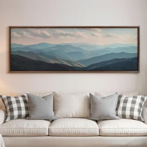May include: A framed landscape painting depicts a mountain range in shades of blue and green under a cloudy sky. The artwork hangs above a cream-colored sofa with gray and checkered pillows.