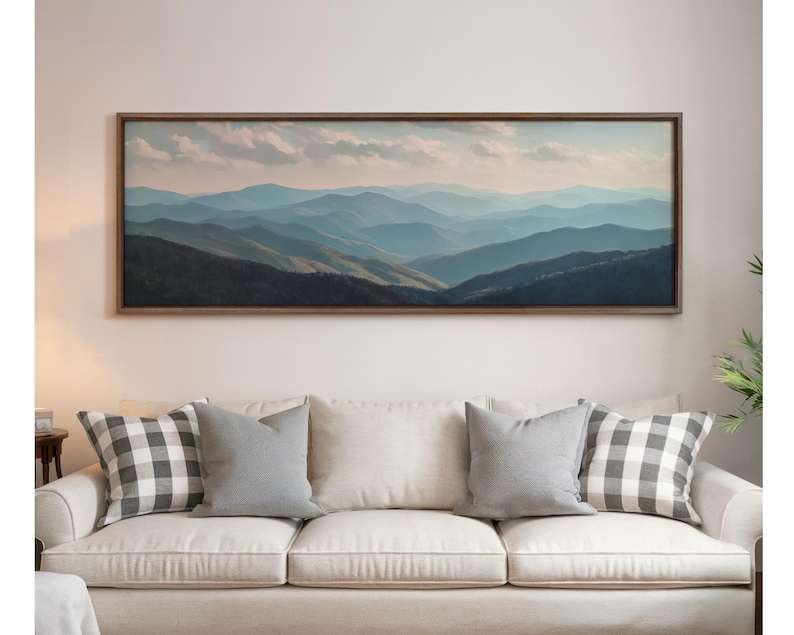 May include: A framed landscape painting depicts a mountain range in shades of blue and green under a cloudy sky. The artwork hangs above a cream-colored sofa with gray and checkered pillows.