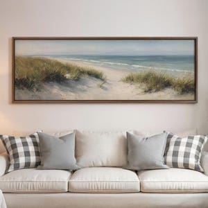 May include: A panoramic landscape painting in a dark wood frame depicting a coastal scene with sand dunes, tall green beach grass, and a calm blue ocean under a soft sky. The artwork is displayed above a white sofa with gray and white pillows.