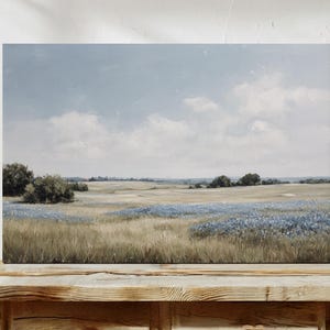 May include: A landscape painting depicting a field of tall, dry grass and blue wildflowers under a cloudy sky. The artwork is displayed on a wooden surface, showcasing a serene, natural scene.