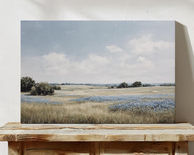 May include: A landscape painting depicting a field of tall, dry grass and blue wildflowers under a cloudy sky. The artwork is displayed on a wooden surface, showcasing a serene, natural scene.