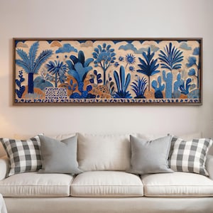 May include: A framed artwork featuring a desert landscape with blue and white plants, cacti, and clouds. The artwork is mounted above a white sofa with gray pillows and checkered throw pillows. The frame is a dark brown color.