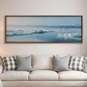 May include: A framed landscape painting depicts a serene arctic scene. The artwork showcases a vast expanse of icy blue water and ice formations under a pale blue sky. The painting is mounted above a cream-colored sofa with gray and checkered pillows.