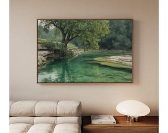 Könnte beinhalten: Ein gerahmtes Landschaftsgemälde zeigt eine ruhige Flussszene. Das Wasser ist ein klares Türkis, das die üppigen grünen Bäume und das Laub entlang der Ufer widerspiegelt. Das Kunstwerk wird über einem beigen Sofa und einem Nachttisch aus Holz mit einer Lampe ausgestellt.