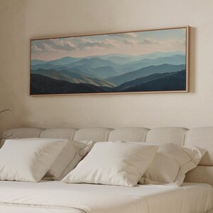 May include: A framed landscape painting of layered blue mountains under a cloudy sky hangs above a bed with white pillows and a cream-colored headboard. The artwork is rectangular with a light wood frame.